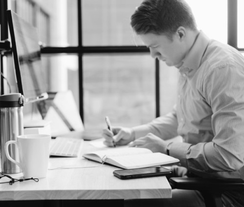 Man sitting at computer desk writing in a notebook.