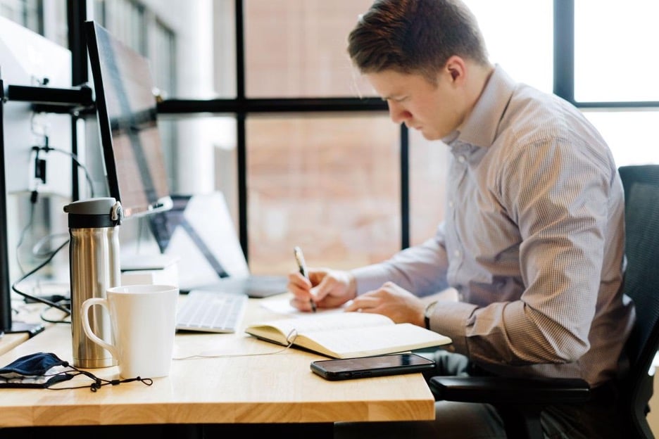 Man sitting at computer desk writing in a notebook.