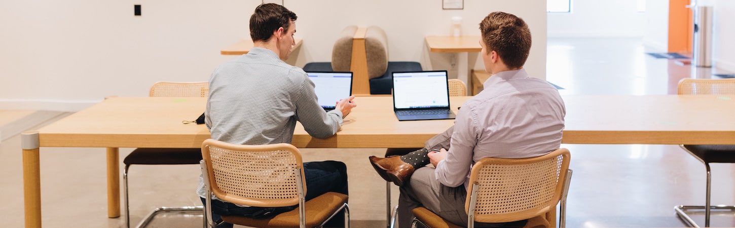 Two men with their backs to the camera sitting at a desk with laptops.